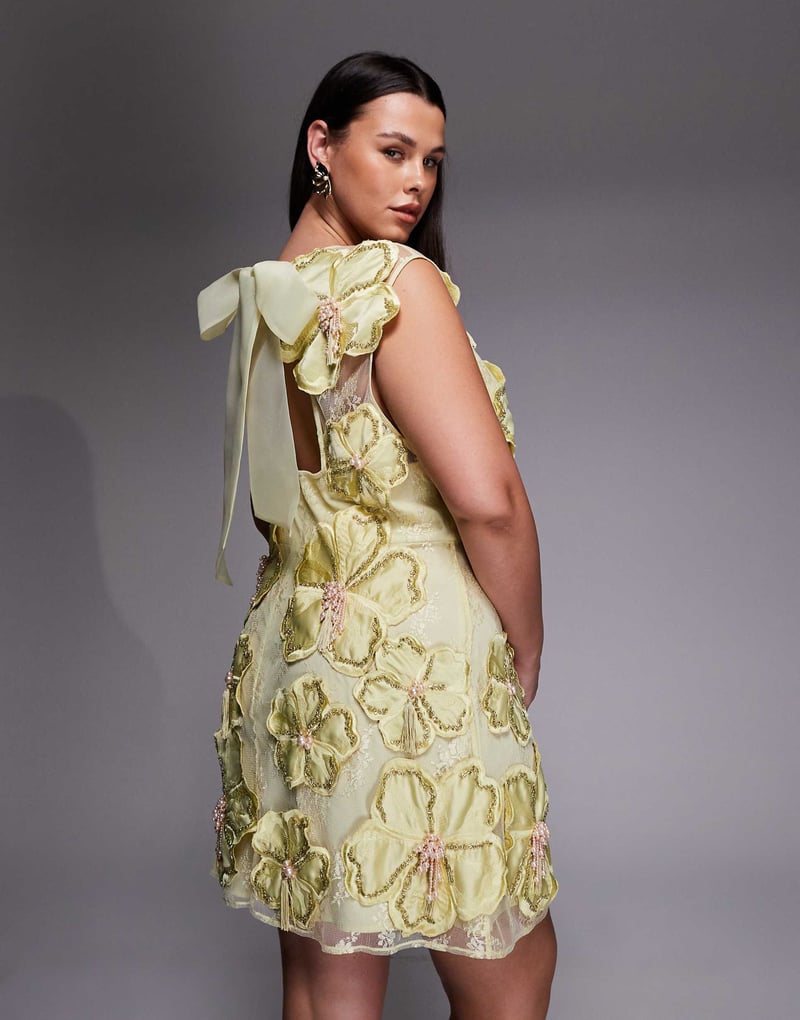a plus size model shows the back of her dress, she is wearing a pale yellow mini dress with 3D florals and oversized bow tie at the neck.