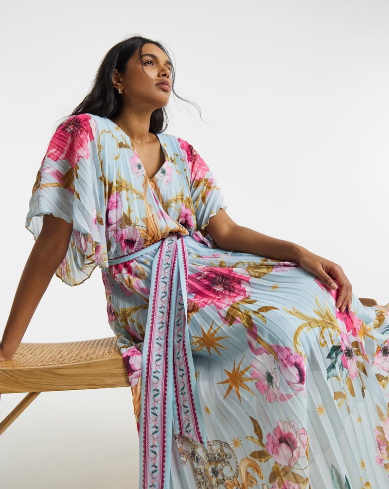 a plus size model sitting down wears a pale blue and bright pink floral wrap dress with belt tie detail.