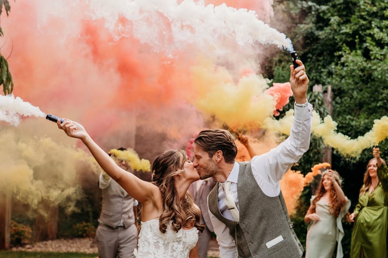 a bride and groom kiss as they hold colourful flares. Their wedding party can be seen holding flares in the background.
