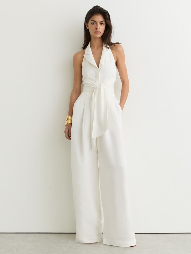 a model wears a cream linen halterneck jumpsuit with pockets and tie waist detail.