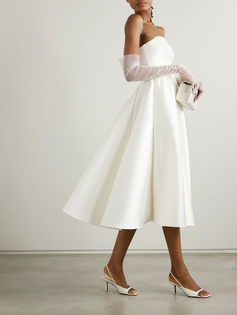 a model wears a strapless pleated mikado satin midi wedding dress with elbow-length chiffon gloves and holds a clutch bag.