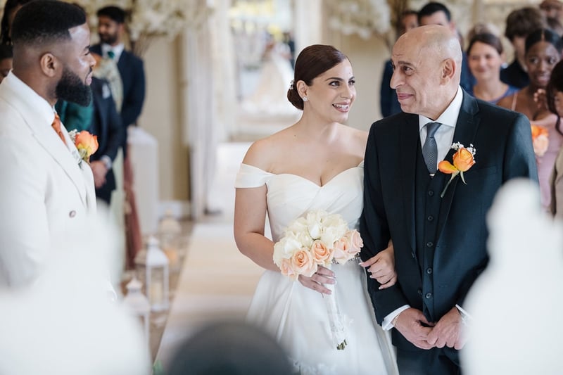 a bride and her father walk down the aisle arm in arm as her groom looks on and smiles.