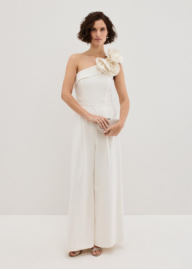 a model wears an ivory one-shoulder jumpsuit with wide leg trousers and corsage detail on the shoulder.