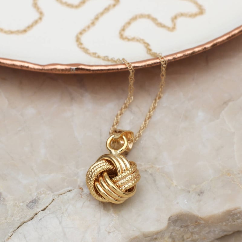 a gold love knot necklace laid across a stone surface next to a ceramic plate
