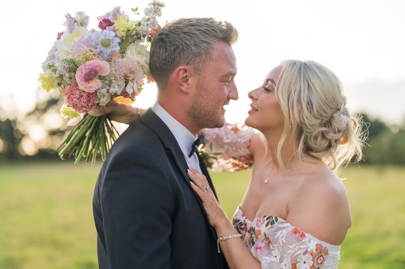 a bride and groom look into each other's eyes. the bride is holding a pastel-coloured bouquet and wearing a wedding dress with coloured floral detailing.