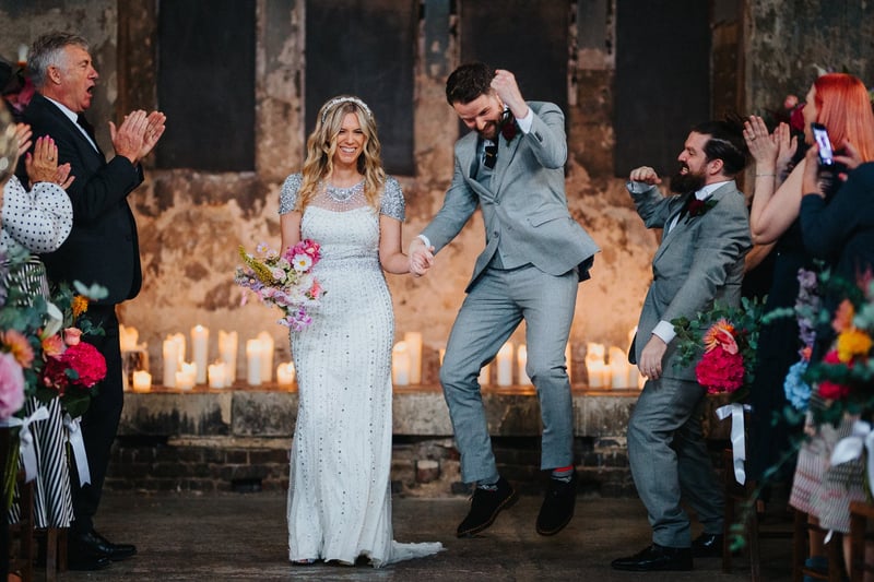 a groom jumps in the air after his ceremony as he holds hand with his bride who is laughing.