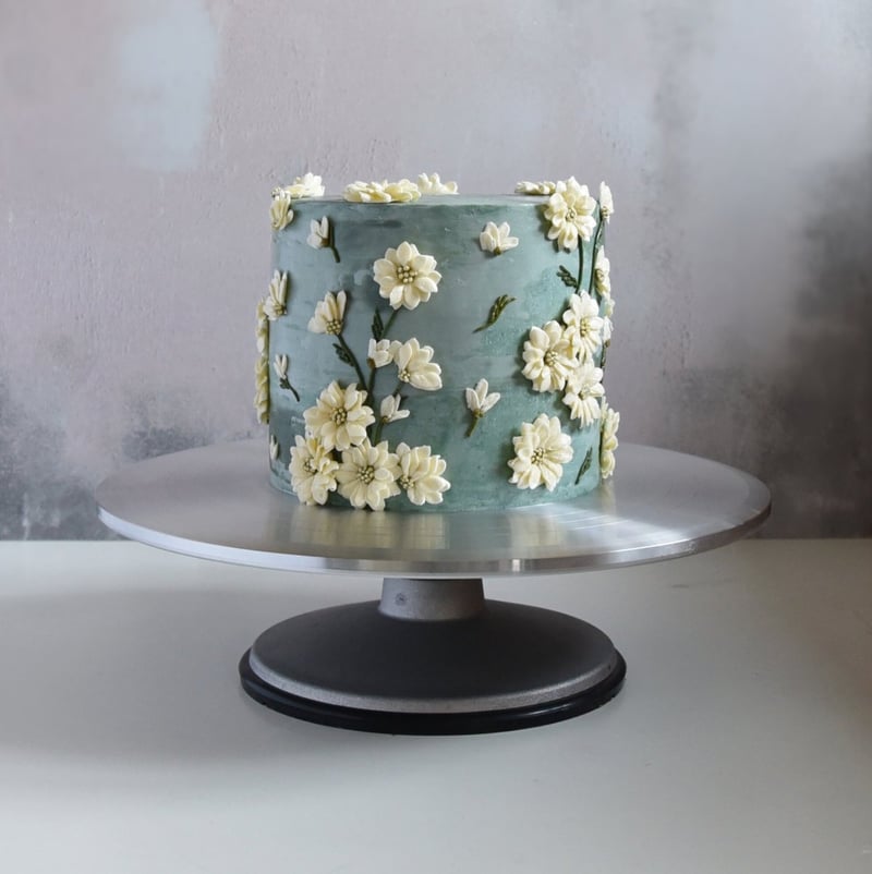 a single tier wedding cake with blue buttercream icing decorated with small piped buttercream daisies.