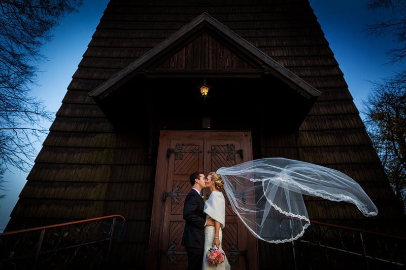 a dark and moody shot of a newlywed bride and groom kissing in front of their wedding venue as the bride's veil blows in the wind.