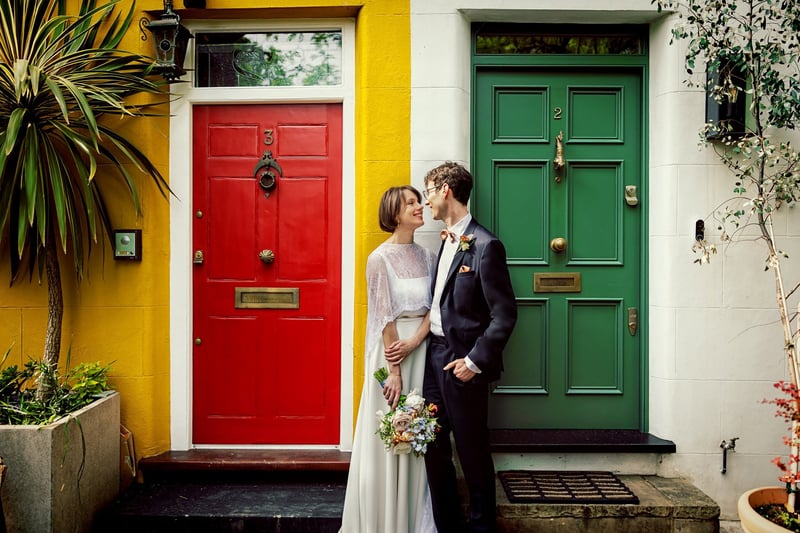 a bride and groom smile at each other as they stand in front of some brightly coloured front doors in London.