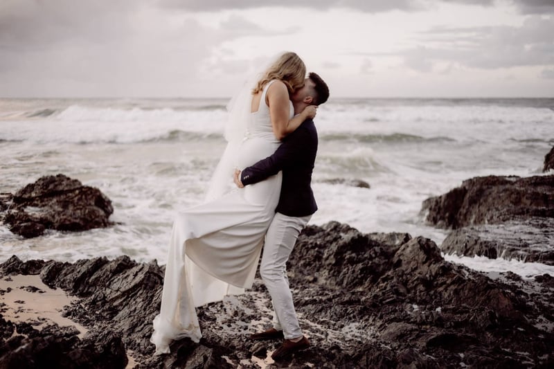 A bride and groom kiss on a beach in australia as waves hit the rocks behind them