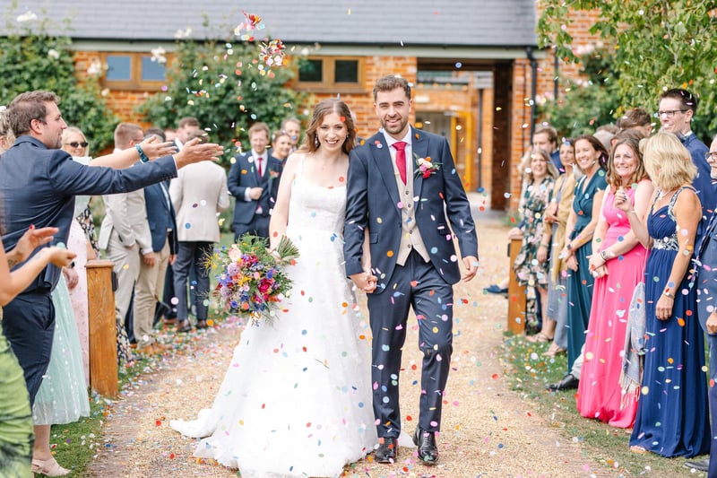 a newlywed bride and groom walk outside their venue as guests throw confetti.