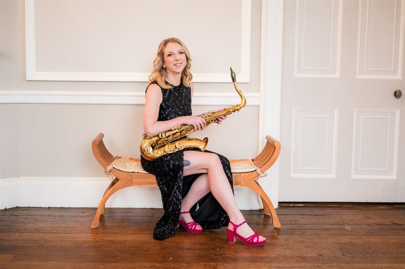 a female saxophonist wearing a black dress and pink shoes smiles as she holds her saxophone.