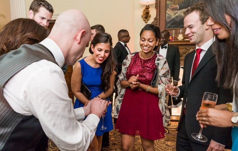 a magician performs a trick at smiling guests in a drinks reception setting.