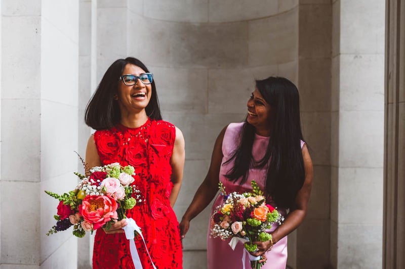 two brides, one wearing red and another wearing pink, laughing as they walk, talk and hold their colourful bouquets.