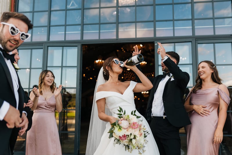 a bride wearing sunglasses drinks champagne from the bottle as guests look on and laugh.