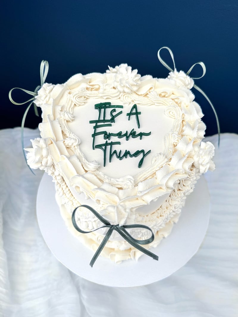a white heart-shaped Lambeth cake with green writing that reads 'it's a forever thing' and green ribbons.