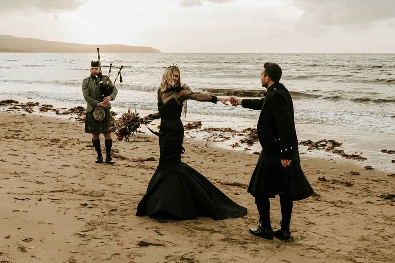 Bride in a black wedding dress and a groom in a kilt dancing on a scottish beach as a bagpiper plays