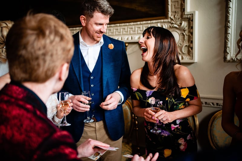 two wedding guests laugh as a wedding magician in a red jacket performs a trick.