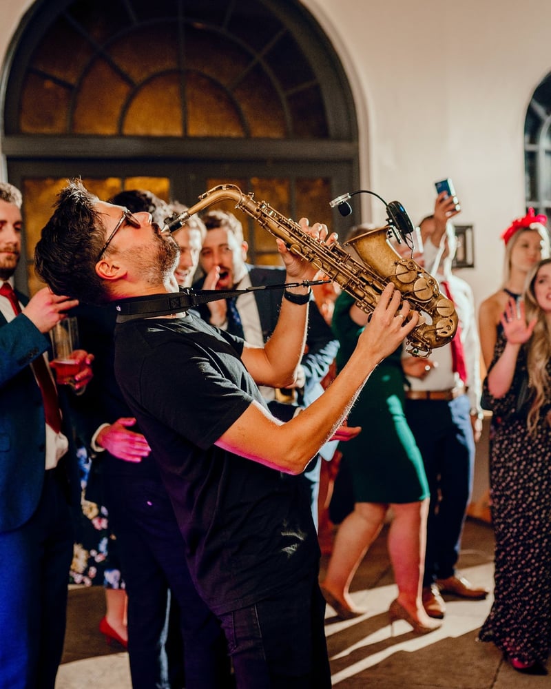 a male saxophonist plays at a wedding reception as guests clap and dance around him.