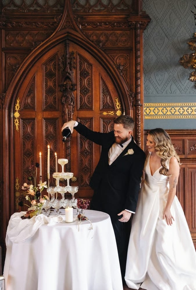 A groom pouring champagne into a tower of glasses as his bride looks on