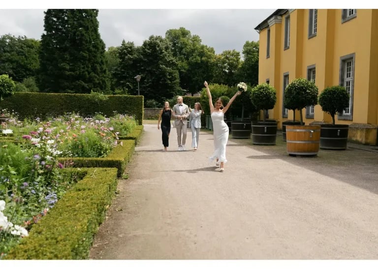 Fröhliche Braut läuft mit erhobenem Brautstrauß durch den Schlossgarten, während Bräutigam und Trauzeuginnen ihr folgen