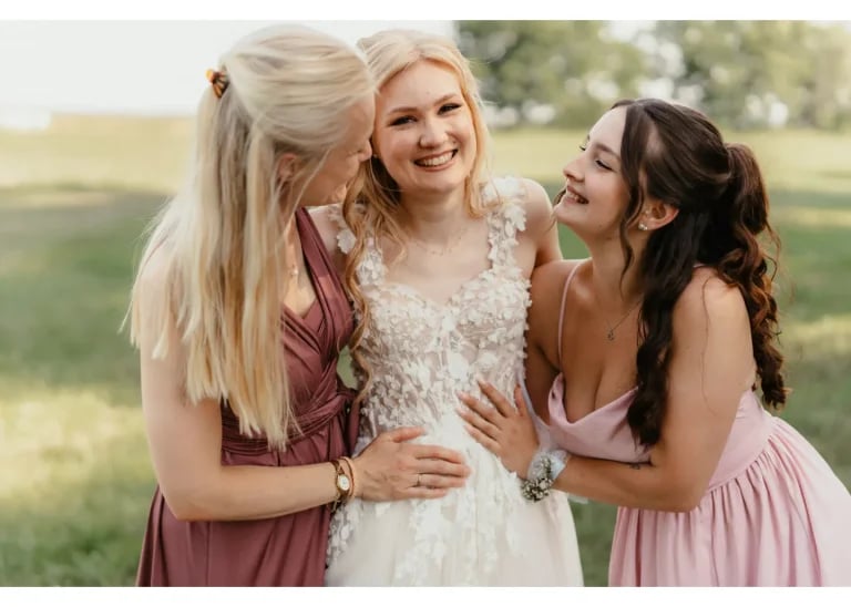 Braut wird von zwei Trauzeuginnen liebevoll umarmt und strahlt vor Glück bei der Hochzeit im Freien