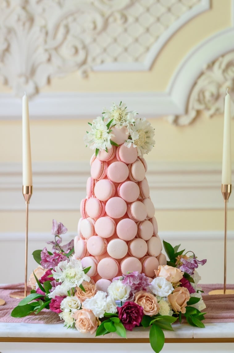 a pink macaron tower surrounded by colourful flowers on a wedding dessert table.