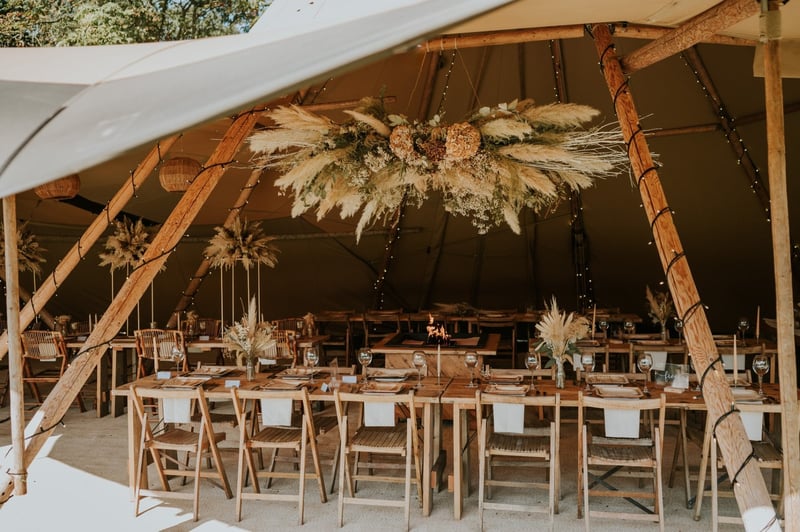 interior of a tipi wedding tent with long wood tables and an overhead floral installation made of pampas grass and dried flowers.