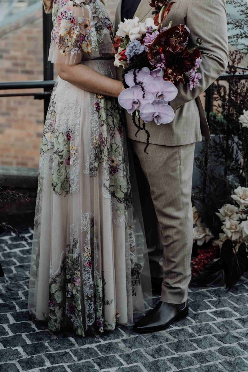 Close up image of an alternative style bride and groom from the shoulders down holding a bouquet of orchids and wearing a colourful floral wedding dress