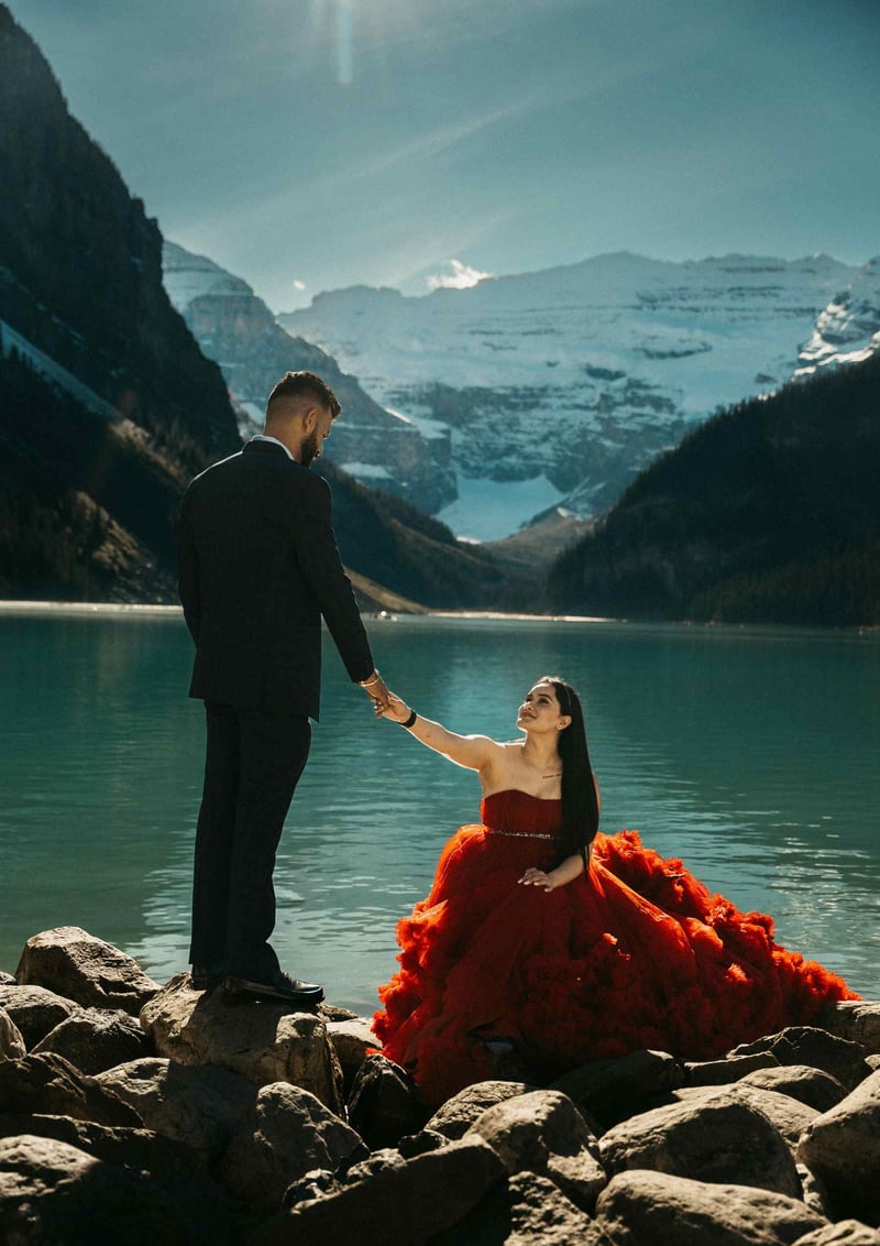 Bride wearing a red wedding dress reaching out to the groom lakeside in the canadian mountains