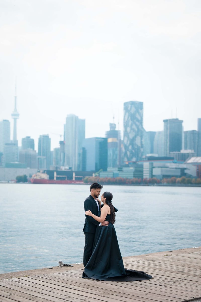 groom with a bride in a black dress embracing at the waterside in toronto with the city skyline behind them