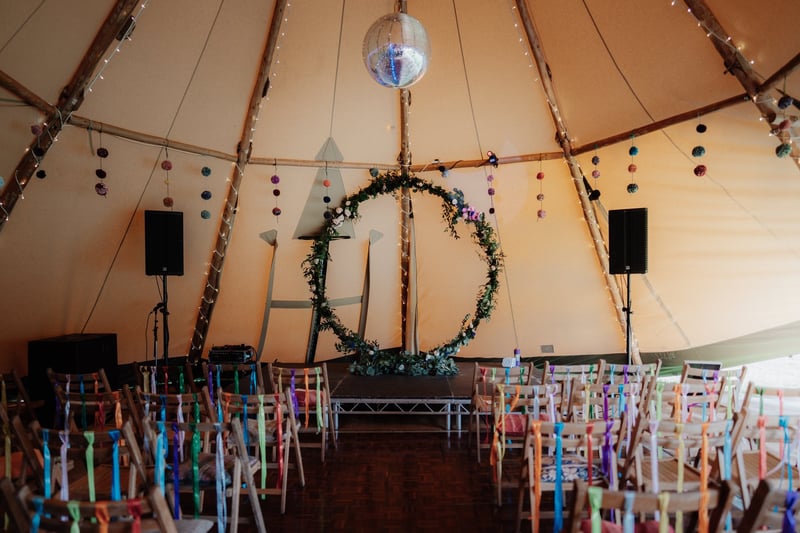 interior of a wedding tipi set up for a ceremony with wooden chairs decorated with colourful ribbons and a disco ball hanging from the ceiling.