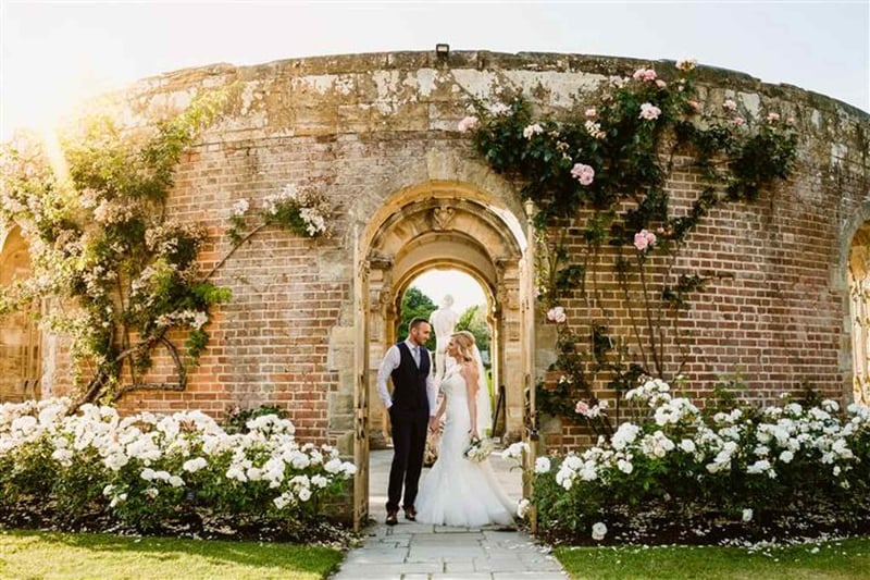 Couple standing in a rose-covered brick archway at a walled garden wedding venue at golden hour