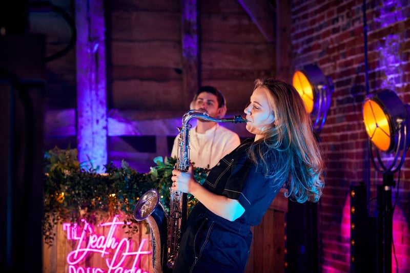 a female saxophonist performs at a wedding reception as a DJ plays in the background.