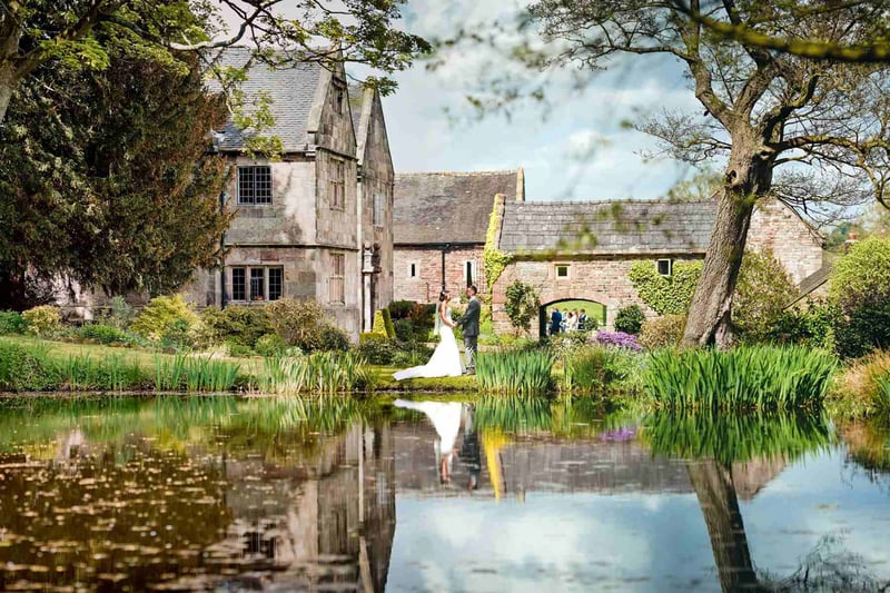  Couple reflected in a lake beside a medieval stone manor house and ivy-clad barn wedding venue