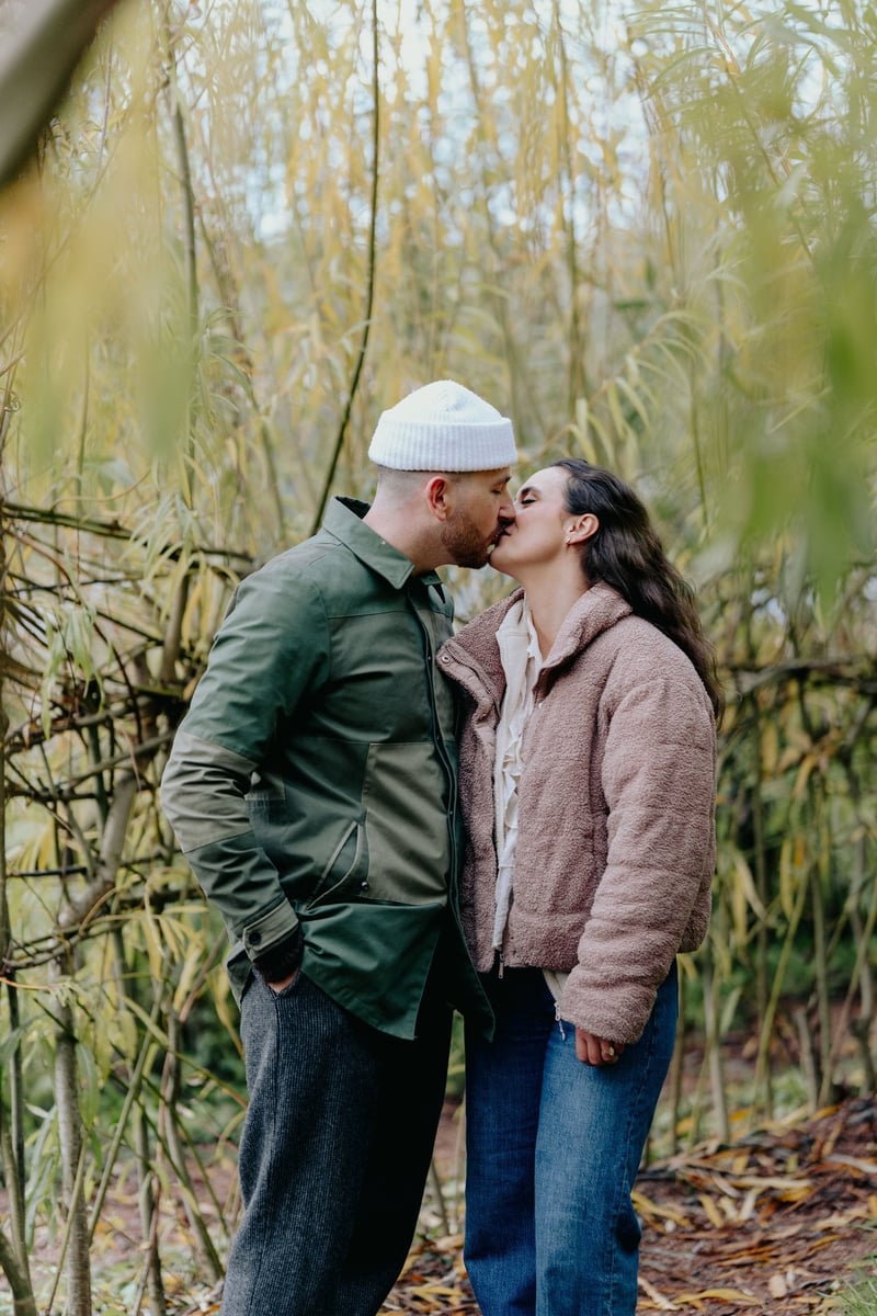 a couple wearing winter clothes kiss in a wooded backdrop.