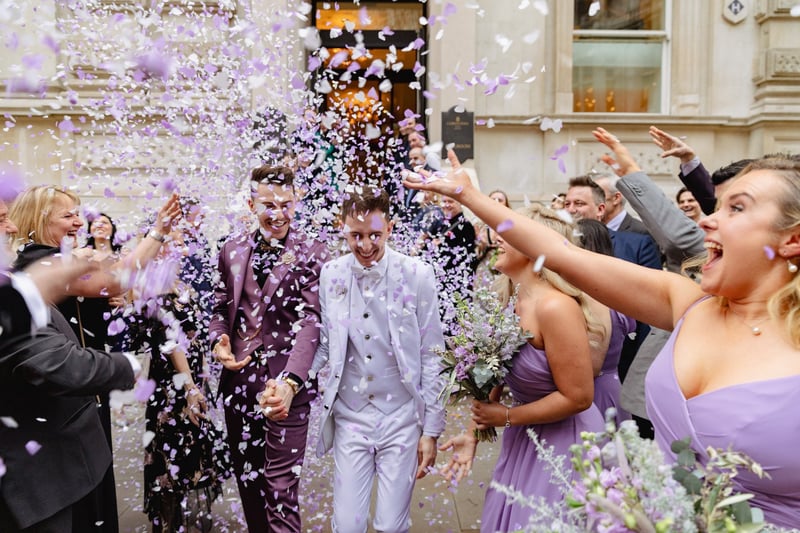 an LGBTQ+ couple depart their ceremony to a shower of purple confetti. They are wearing purple suits.