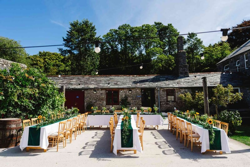 Outdoor wedding reception setup at a rustic Cornish stone farm venue with sunflower table garlands and festoon lights