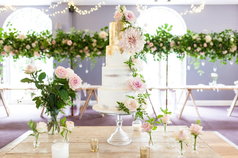 a four tier white buttercream wedding cake with gold leaf and pink floral decorations on a table with flowers.