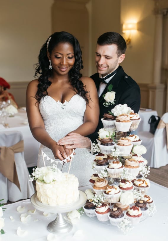 A bride and groom cutting into a single tier wedding cake next to a wedding cupcake tower