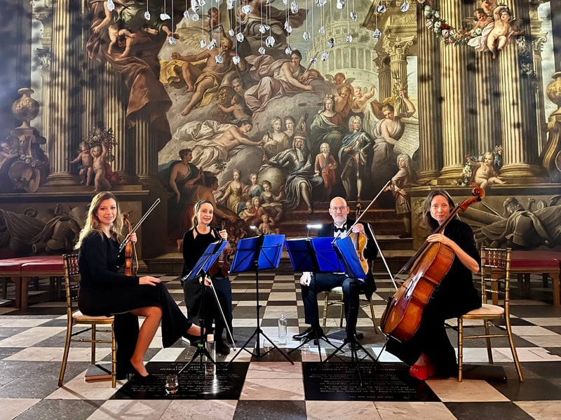 a string quartet smile at the camera as they are seated to perform in a grand building.
