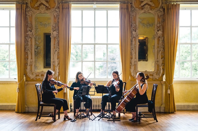 a string quartet dressed in black perform in front of a window in a country house wedding venue.