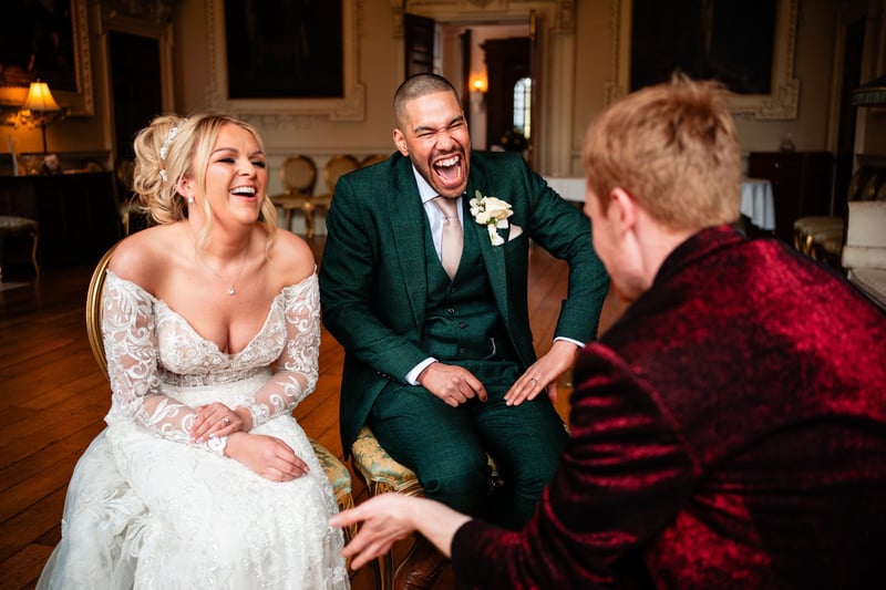 a magician in a red jacket speaks to a laughing bride and groom who are seated.