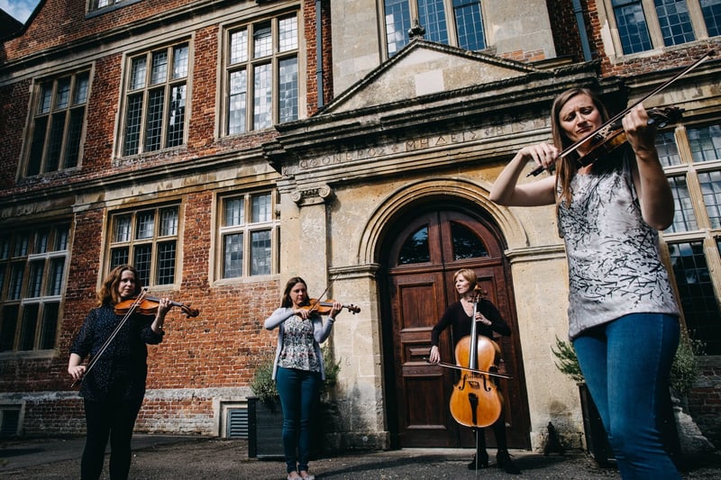 a group of four female musicians perform with their string instruments outside a country house.