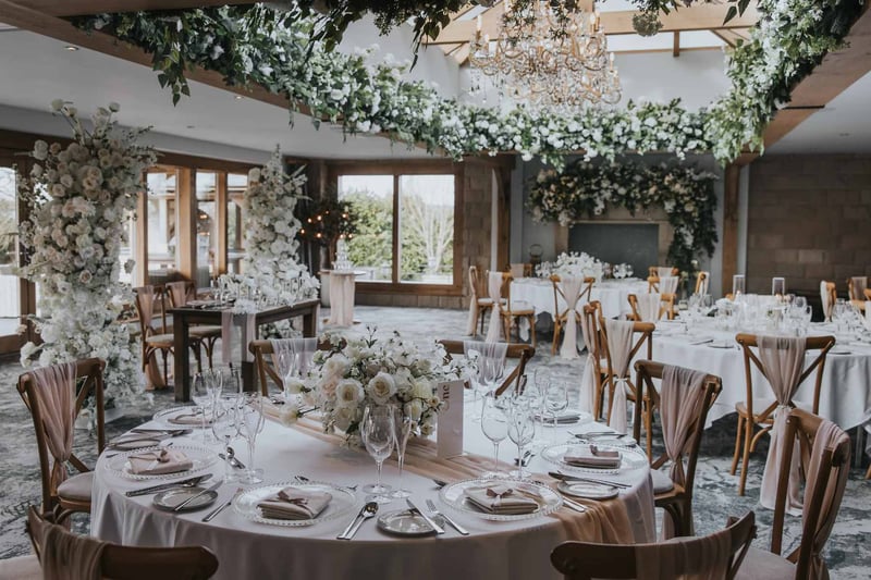 Elegant wedding reception room with ceiling floral installation, crystal chandeliers, and white rose centrepieces