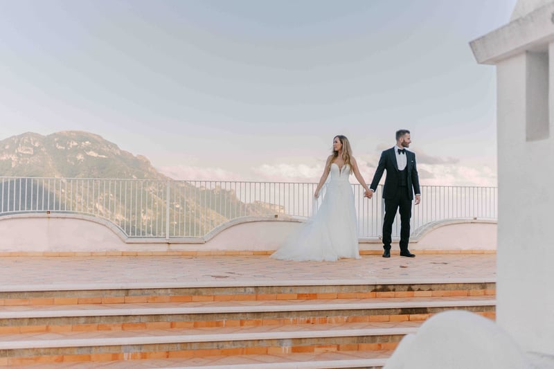 a couple pose against the backdrop of sea and mountains in their wedding attire.