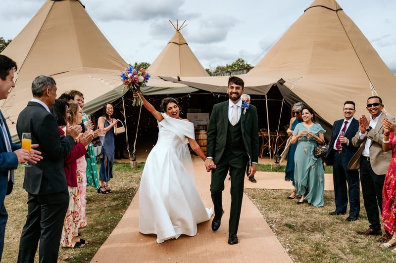 a newlywed bride and groom walk out of their tipi wedding tent as guests clap.