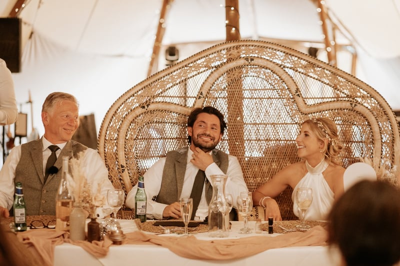 a bride and groom smile as they sit at the top table with the father of the bride.