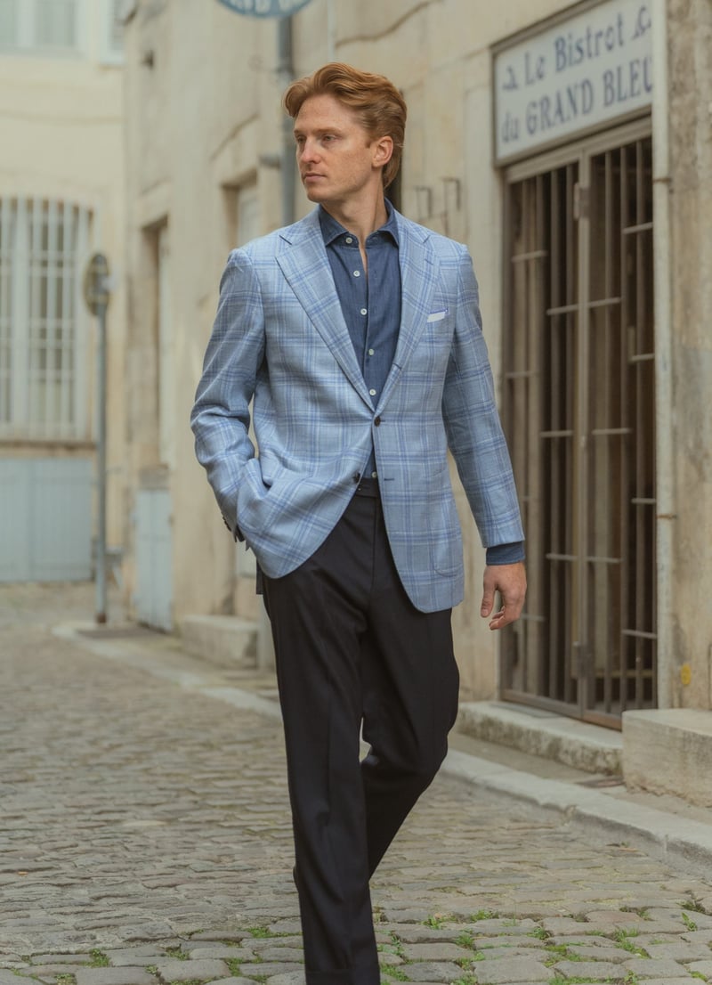 a man walks down a European side street wearing navy trousers and a light blue checked tailored jacket.