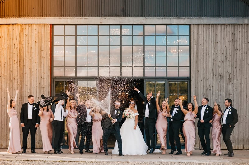 a wedding party cheer and wave their arms in the air as they pop champagne.